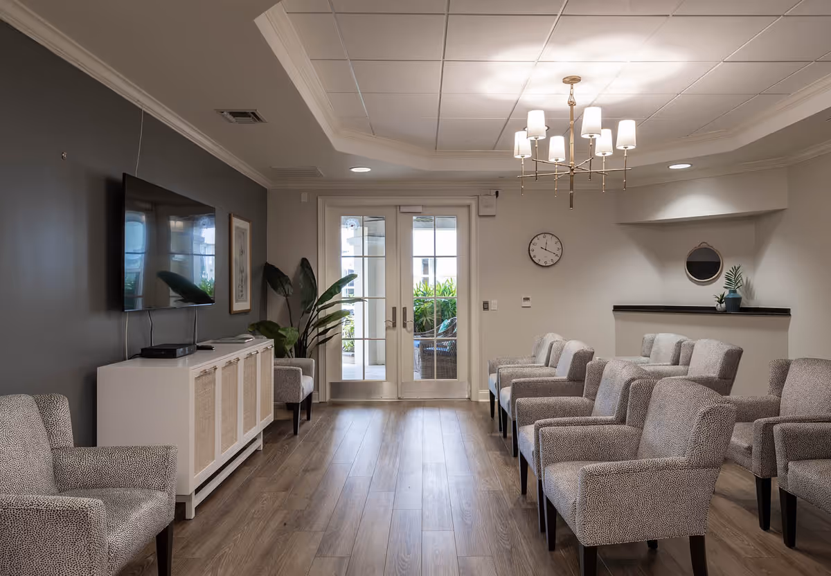 A well-lit waiting or common area with multiple patterned armchairs arranged in rows facing a wall-mounted flat screen TV. The room has wooden flooring, a modern chandelier, a clock on the wall, a small plant on a shelf, and double glass doors leading outside with greenery visible through the doors.