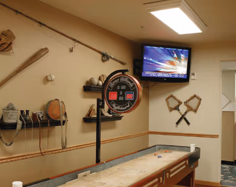 Interior room with a shuffleboard table in the foreground, a mounted television on the wall showing a sports channel, and various vintage sports equipment including baseball bats, gloves, and balls displayed on shelves and mounted on the walls.