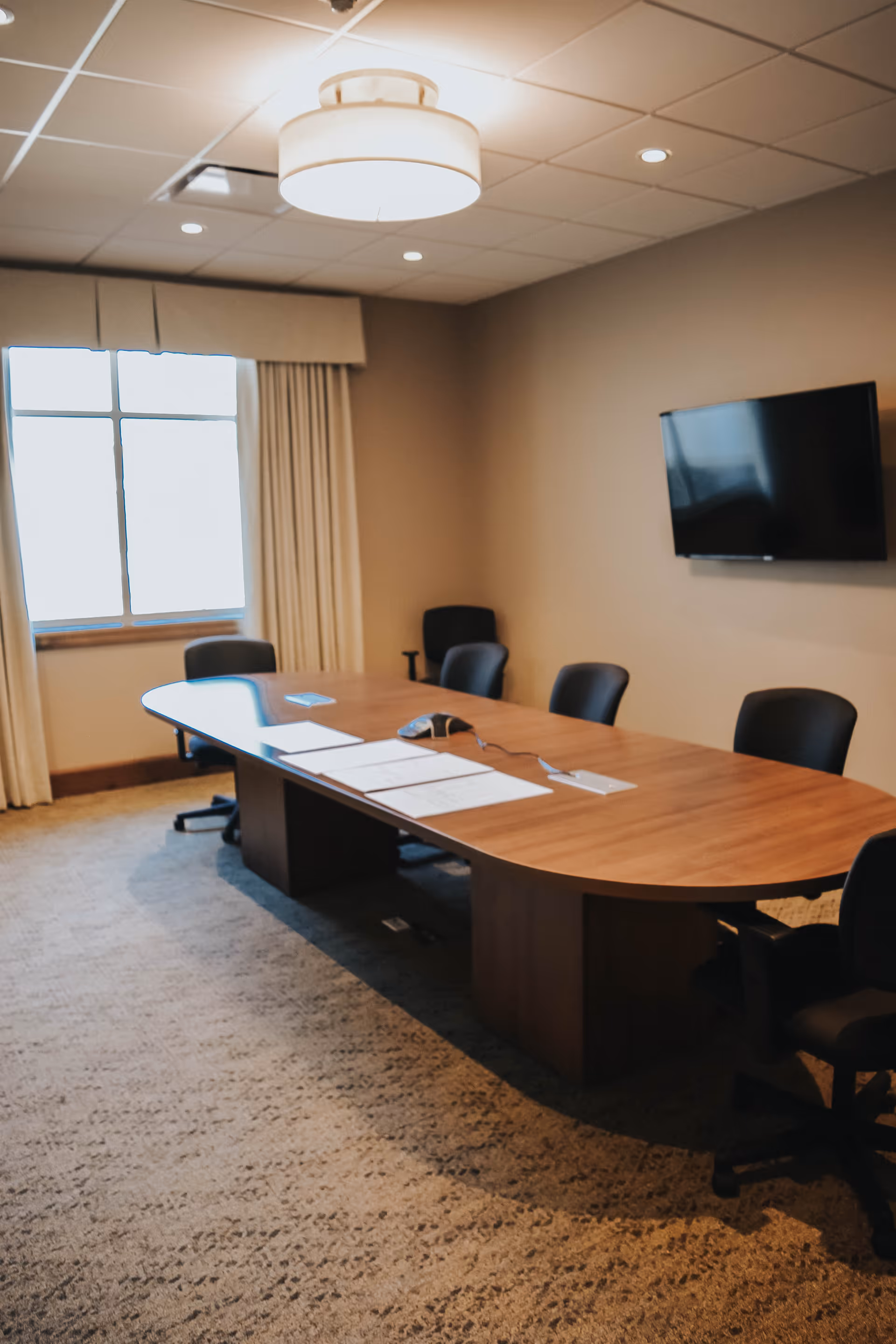 A conference room with a long wooden table surrounded by black office chairs. There are papers and a conference phone on the table. A large window with curtains is on one side, and a flat-screen TV is mounted on the wall. The room has a beige carpet and a ceiling light fixture.