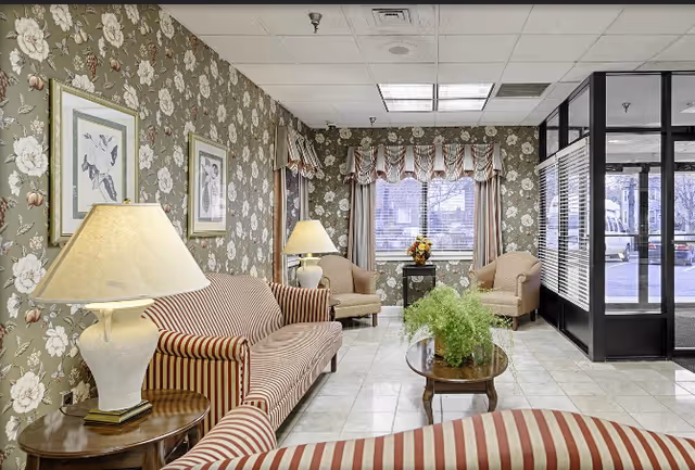 A cozy living room area with floral wallpaper, striped upholstered sofas and armchairs, two table lamps on wooden side tables, a coffee table with a green plant, and a window with patterned curtains letting in natural light. The room has a tiled floor and a glass door leading outside.