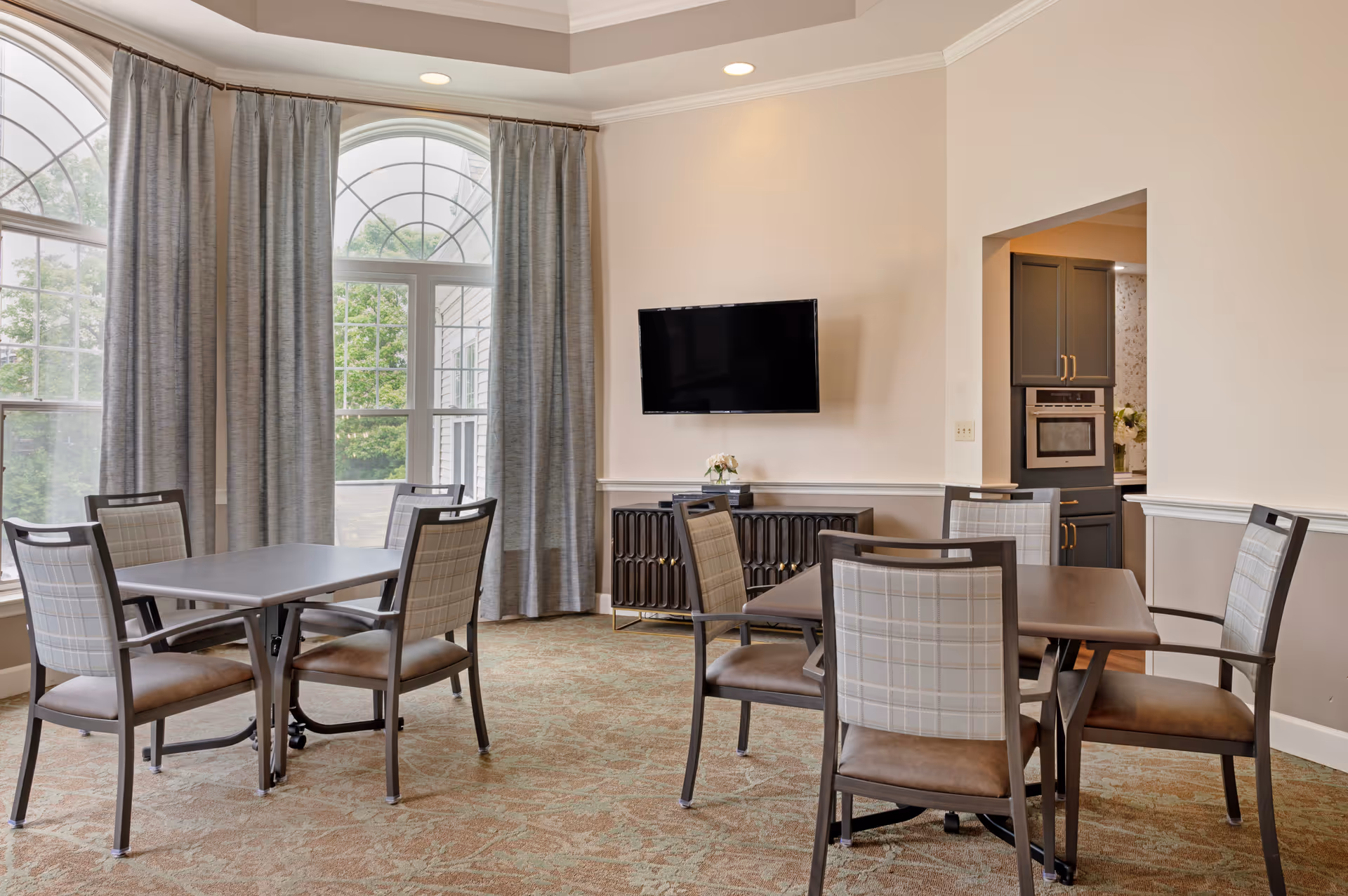 A bright dining area with two square tables, each surrounded by four chairs with plaid upholstery. Large arched windows with gray curtains allow natural light to fill the room. A flat-screen TV is mounted on the beige wall above a dark sideboard. The room has a patterned carpet and an open doorway leading to a kitchen area with gray cabinets.