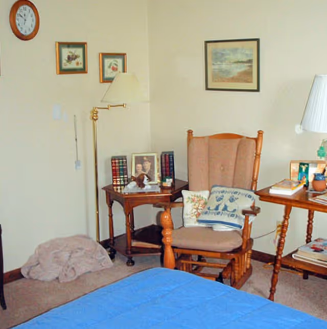 A cozy room corner featuring a wooden rocking chair with cushions, a small wooden side table with framed photos and books, a floor lamp, a wall clock, and framed artwork on the walls. A blue bedspread is partially visible in the foreground.