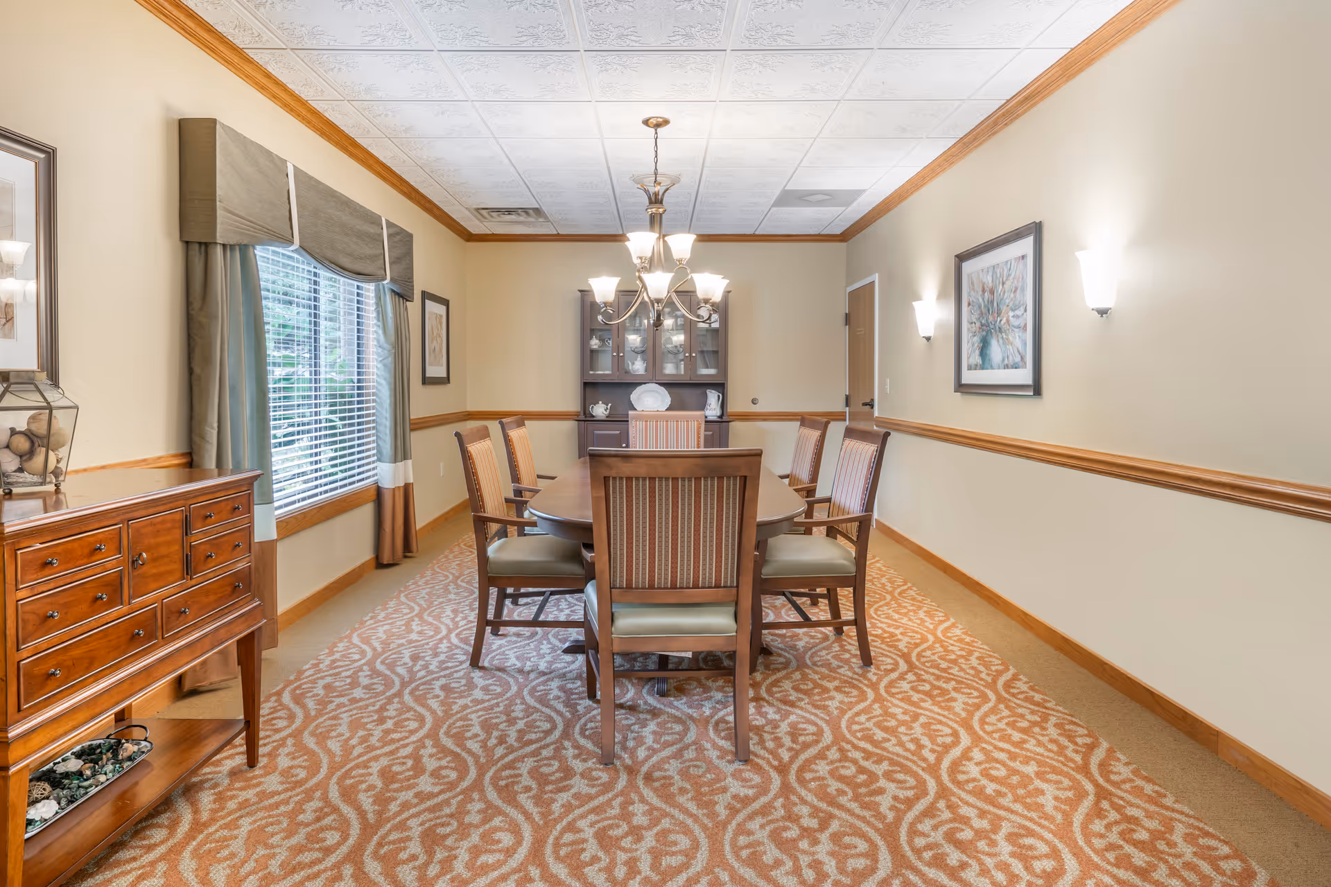 A dining room with a long wooden table surrounded by eight chairs with striped upholstery. The room has beige walls with wooden trim, a patterned carpet, a window with green curtains, a wooden sideboard on the left, a chandelier hanging above the table, and a cabinet with dishes against the far wall.