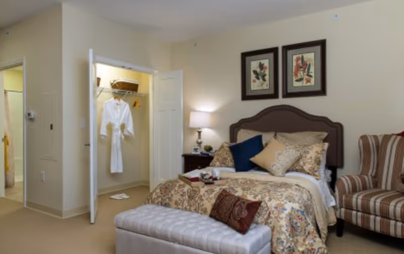 A cozy bedroom with a bed adorned with multiple pillows and a floral patterned comforter. There is a padded bench at the foot of the bed, a striped armchair to the right, and a nightstand with a lamp and decorative items. Two framed floral artworks hang above the headboard. An open closet with a white robe hanging inside is visible, and a bathroom entrance can be seen in the background.