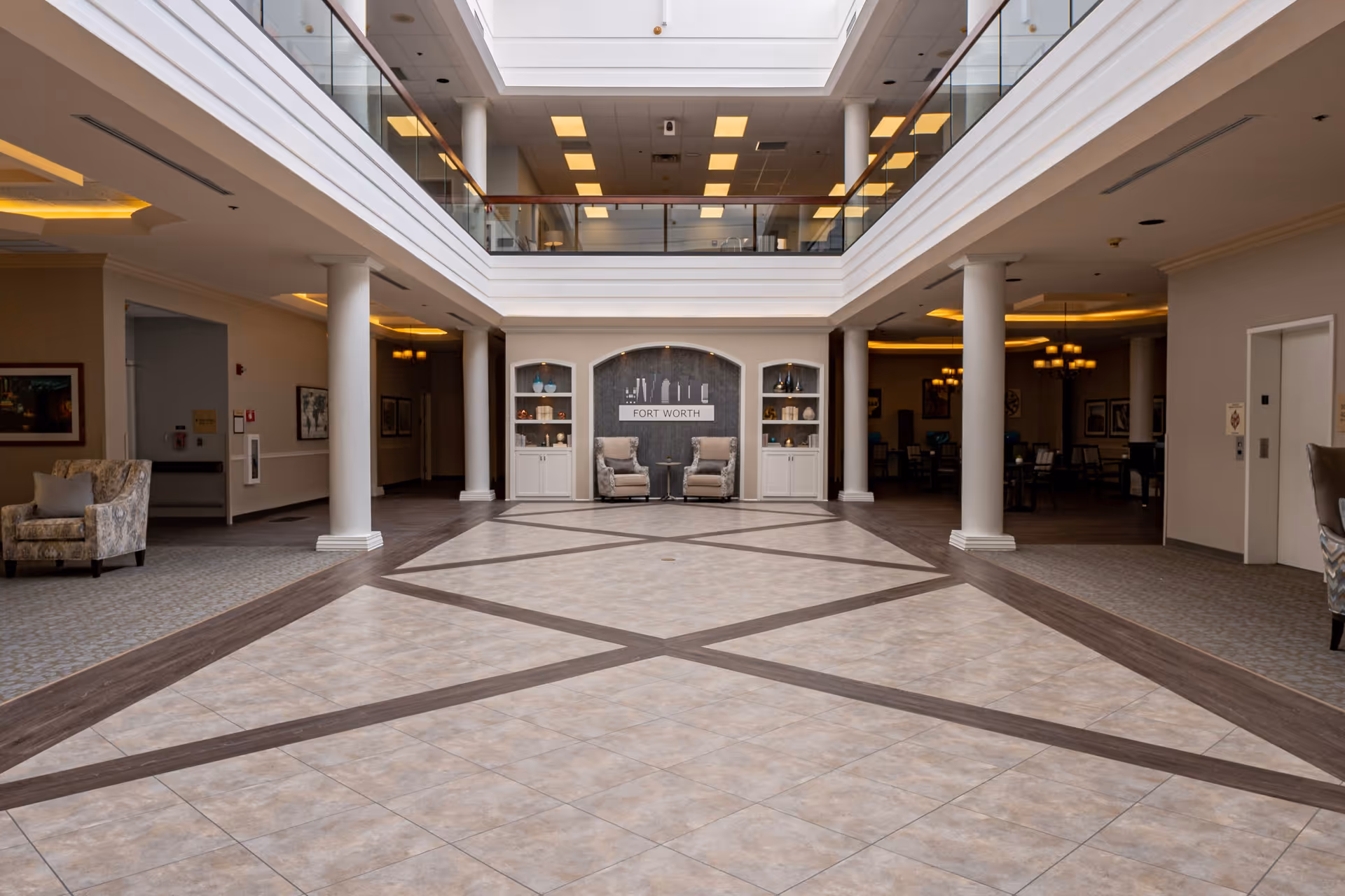 Spacious and well-lit interior lobby area of a senior living facility with tiled floor featuring a geometric pattern, white columns, two armchairs placed against a wall with built-in shelves, and a second-floor balcony with glass railing overlooking the space.