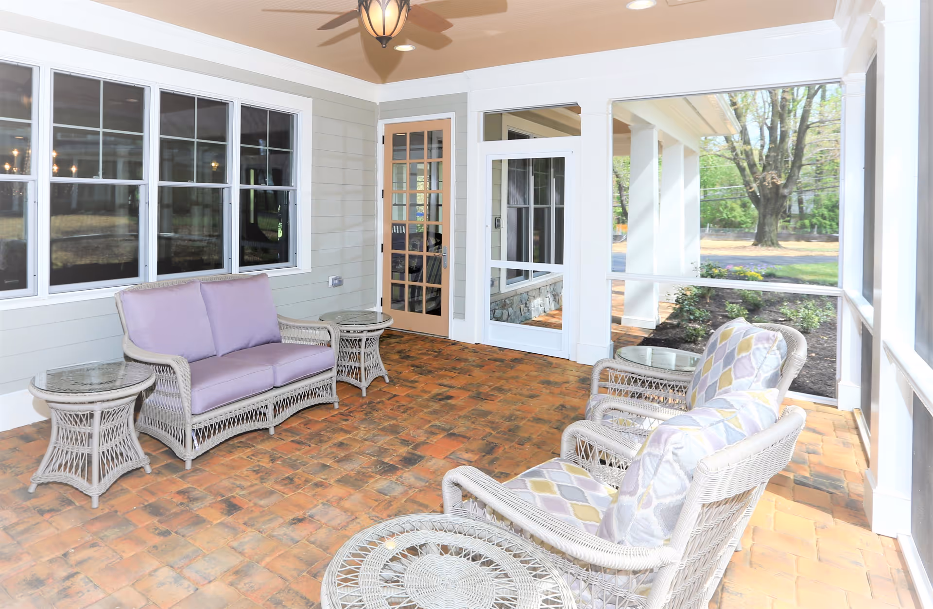 A bright and airy screened-in porch with brick flooring, featuring wicker furniture including a loveseat with purple cushions, two armchairs with patterned cushions, and three glass-topped wicker side tables. The porch is adjacent to a building with large windows and a door leading inside, and overlooks a garden area with trees and shrubs.