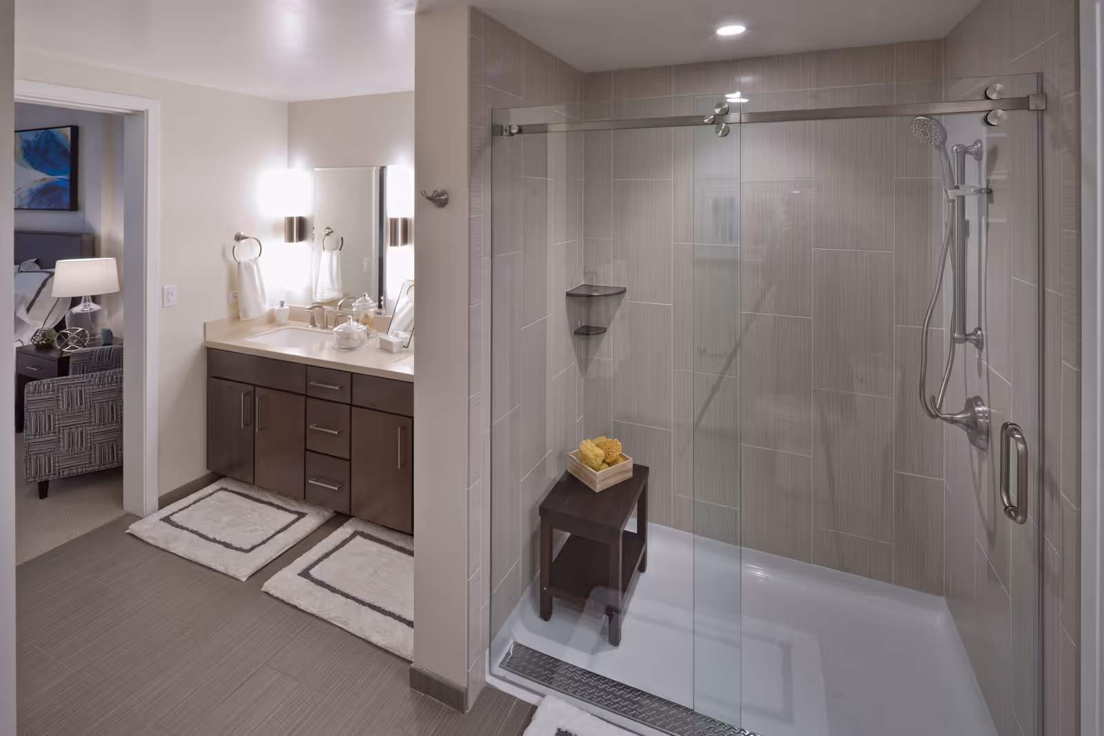 Modern bathroom with a glass-enclosed shower featuring a handheld showerhead and a small wooden stool with sponges. The bathroom has a vanity with a sink, mirror, and two wall-mounted lights. A doorway leads to a bedroom with a bed, nightstand, lamp, and chair visible.