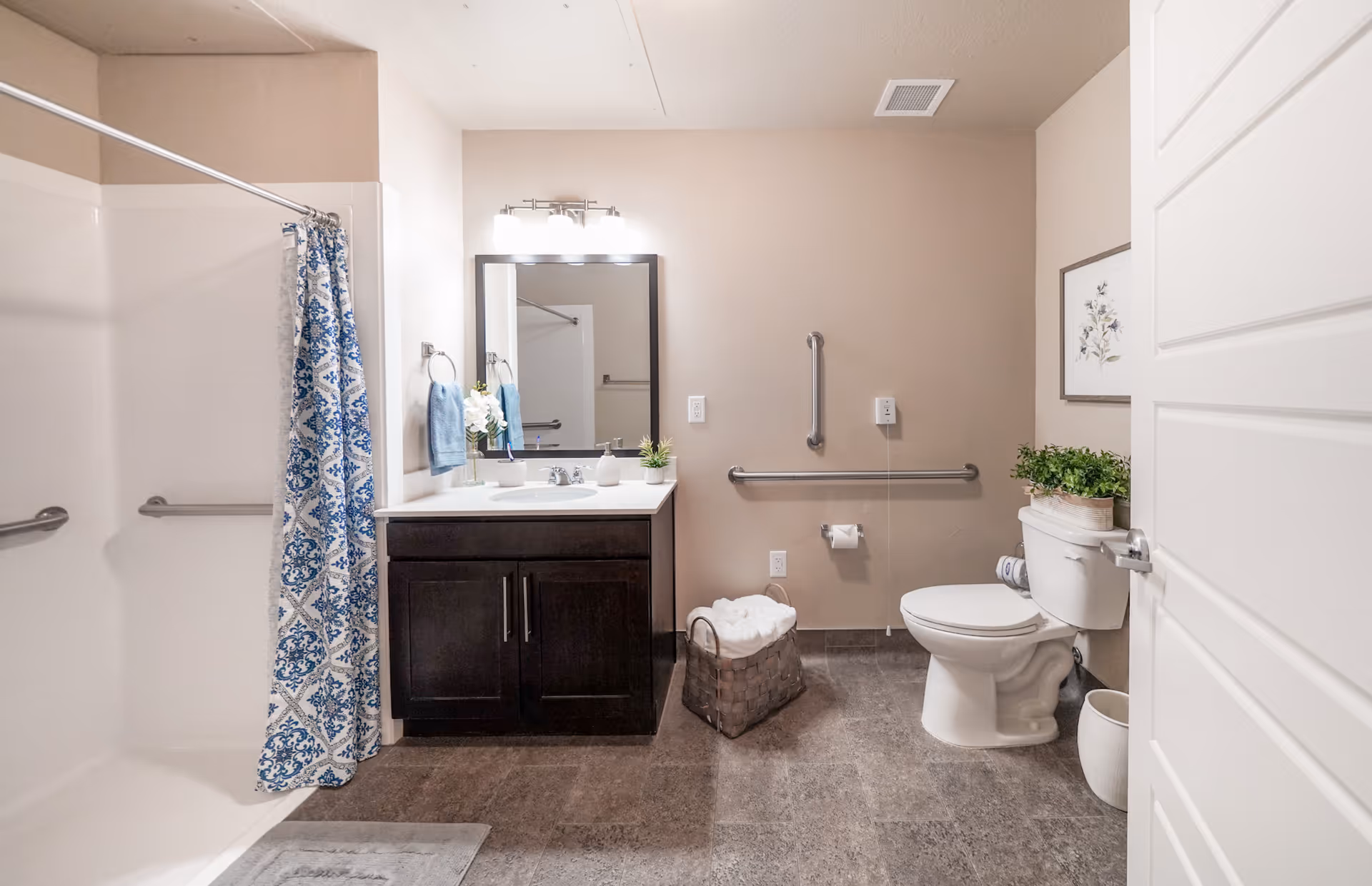 Well-lit accessible bathroom with a vanity and mirror, walk-in shower with a blue patterned curtain, and a toilet with grab bars.