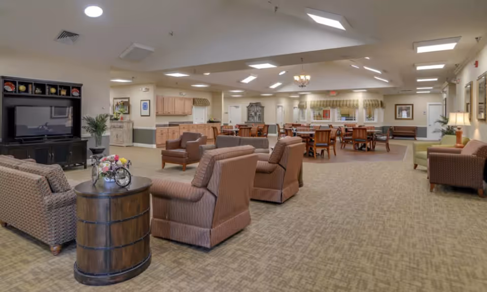 Spacious senior living common room with upholstered chairs and sofas, a TV cabinet, dining tables and a kitchenette under recessed lighting.
