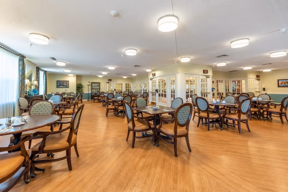 A spacious dining room in a senior living facility with multiple wooden tables and chairs arranged neatly. The room has large windows with curtains allowing natural light to fill the space, wood flooring, and soft ceiling lights. The walls are painted in light colors with some artwork and plants for decoration.