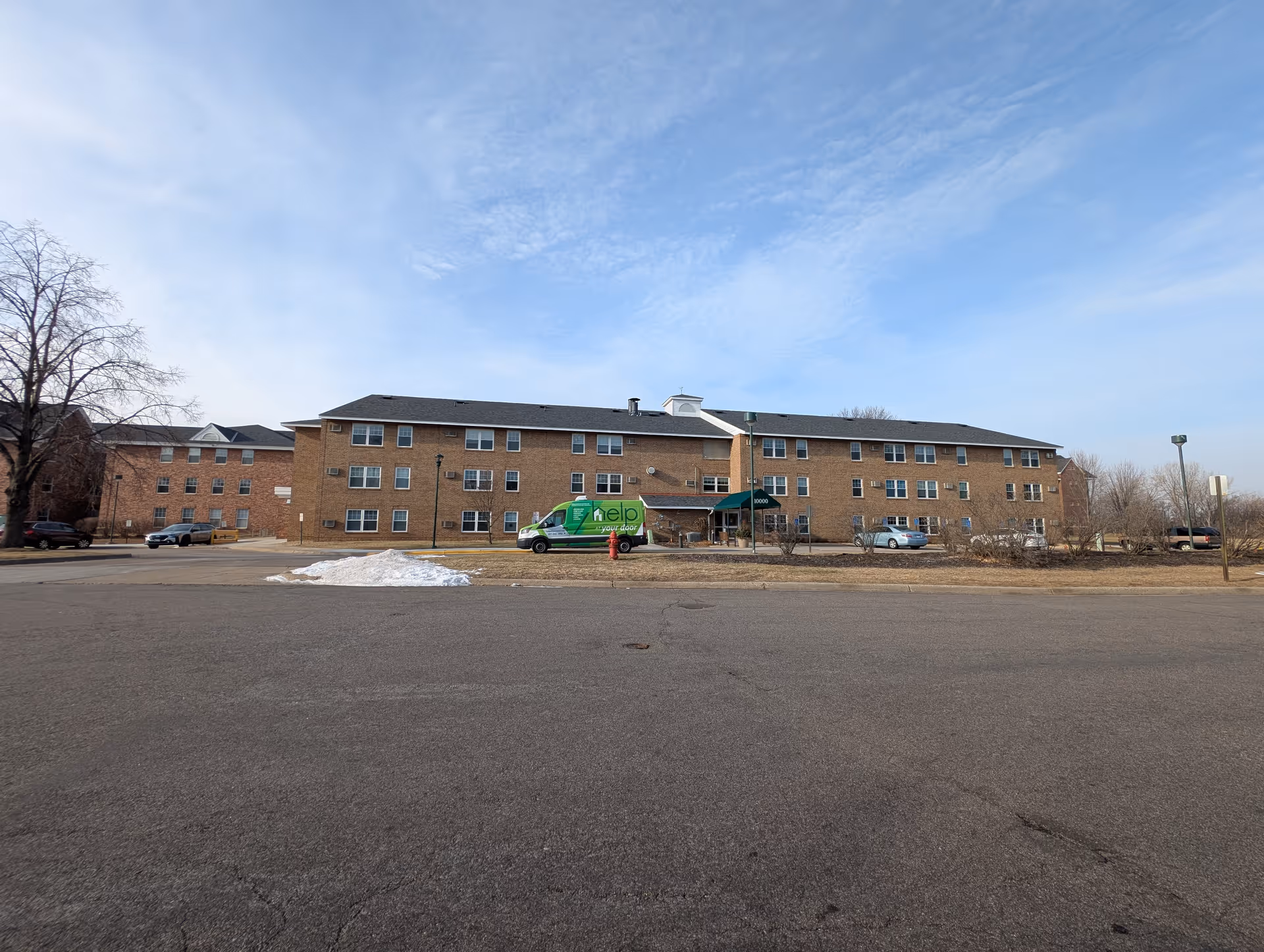 Wide exterior view of a three-story brick building with multiple windows and a green awning over the entrance. A green van with the word 'help' is parked in front, along with a few other cars. There is a small patch of snow on the grass near the parking lot, and leafless trees are visible on either side of the building under a partly cloudy sky.