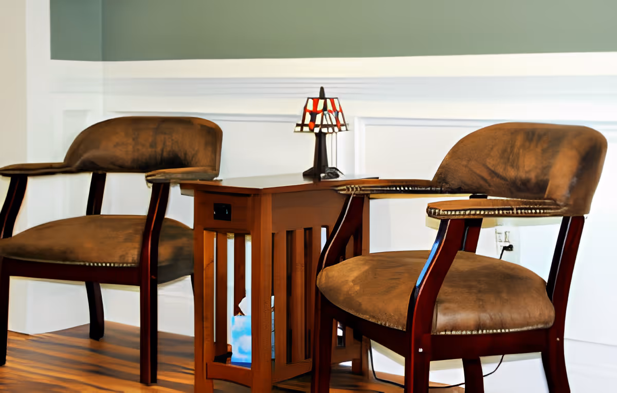 Two brown cushioned wooden armchairs with a small wooden side table between them, featuring a small decorative lamp on top, set against a white paneled wall and wooden floor.