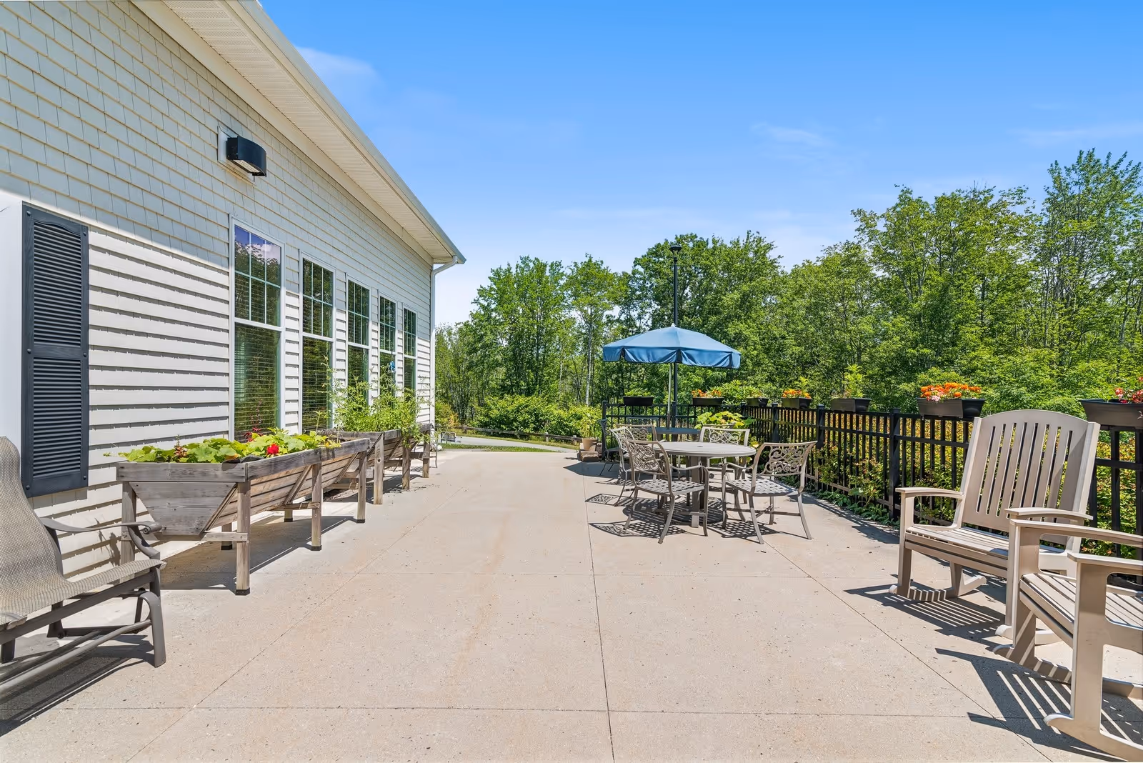 Outdoor patio area at The Enclave of Scarborough with several chairs, a round table with an umbrella, planter boxes with greenery, and a black fence. Trees and blue sky are visible in the background.
