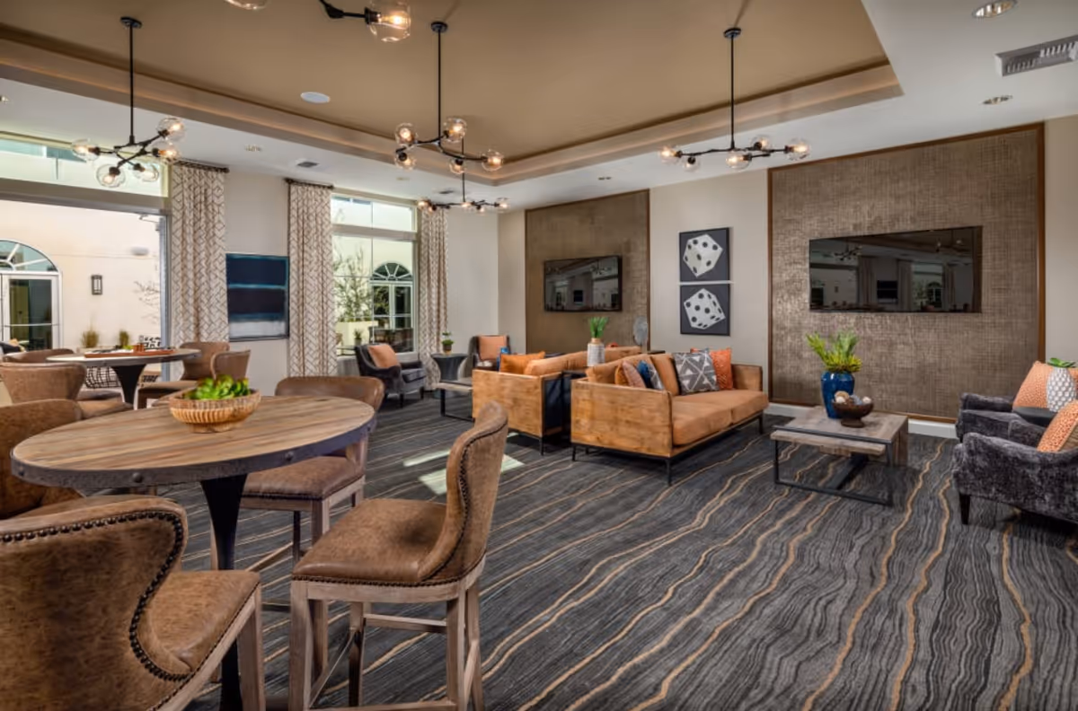 A spacious and well-lit living room area with modern decor featuring two wooden sofas with cushions, two gray armchairs, and a wooden coffee table. The room has patterned carpet flooring, two large wall-mounted flat-screen TVs, and decorative wall art with dice images. There are also round wooden tables with brown upholstered chairs, large windows with patterned curtains, and contemporary ceiling light fixtures.