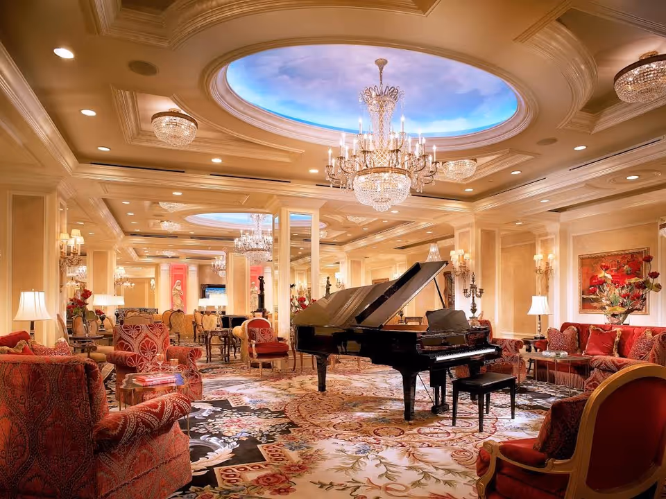 Luxurious living room with a grand piano in the center, surrounded by elegant red upholstered chairs and sofas. The room features ornate chandeliers, a decorative ceiling with a sky mural, and a richly patterned carpet. Warm lighting and classical decor create an inviting atmosphere.