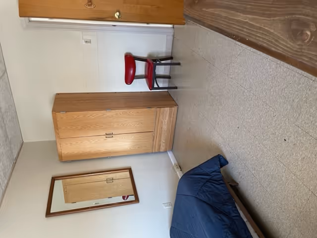 A simple bedroom with a bed covered in a dark blue blanket on the left, a wooden wardrobe with two doors and two drawers in the center against a white wall, a red chair with black legs next to the wardrobe, a wooden door on the right, and a rectangular mirror hanging on the wall above an electrical outlet.