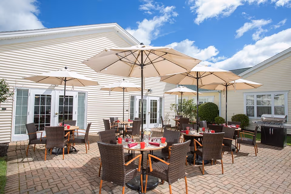 Outdoor patio area with several round tables and wicker chairs, each table shaded by large beige umbrellas. Tables are set with red cups, napkins, and glassware. The patio is paved with bricks and surrounded by light-colored building walls with multiple windows and doors. A barbecue grill is visible on the right side near some bushes.