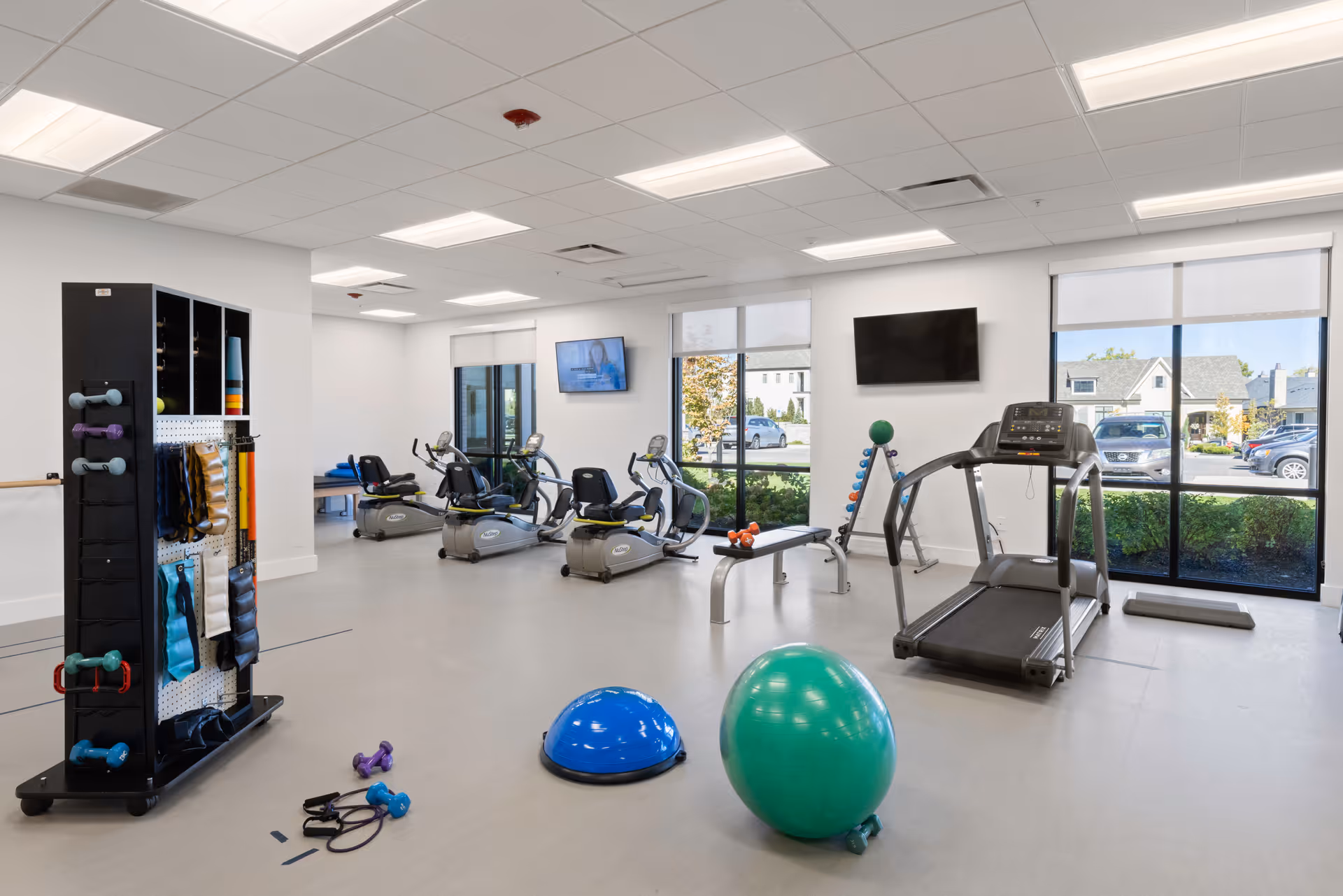 A bright fitness room in a senior living facility with exercise equipment including a treadmill, recumbent bikes, a bench with dumbbells, a rack with various exercise accessories, and large windows letting in natural light.