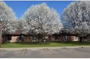 Exterior view of a single-story brick building partially obscured by three large trees with white blossoms, with a parking lot in the foreground.