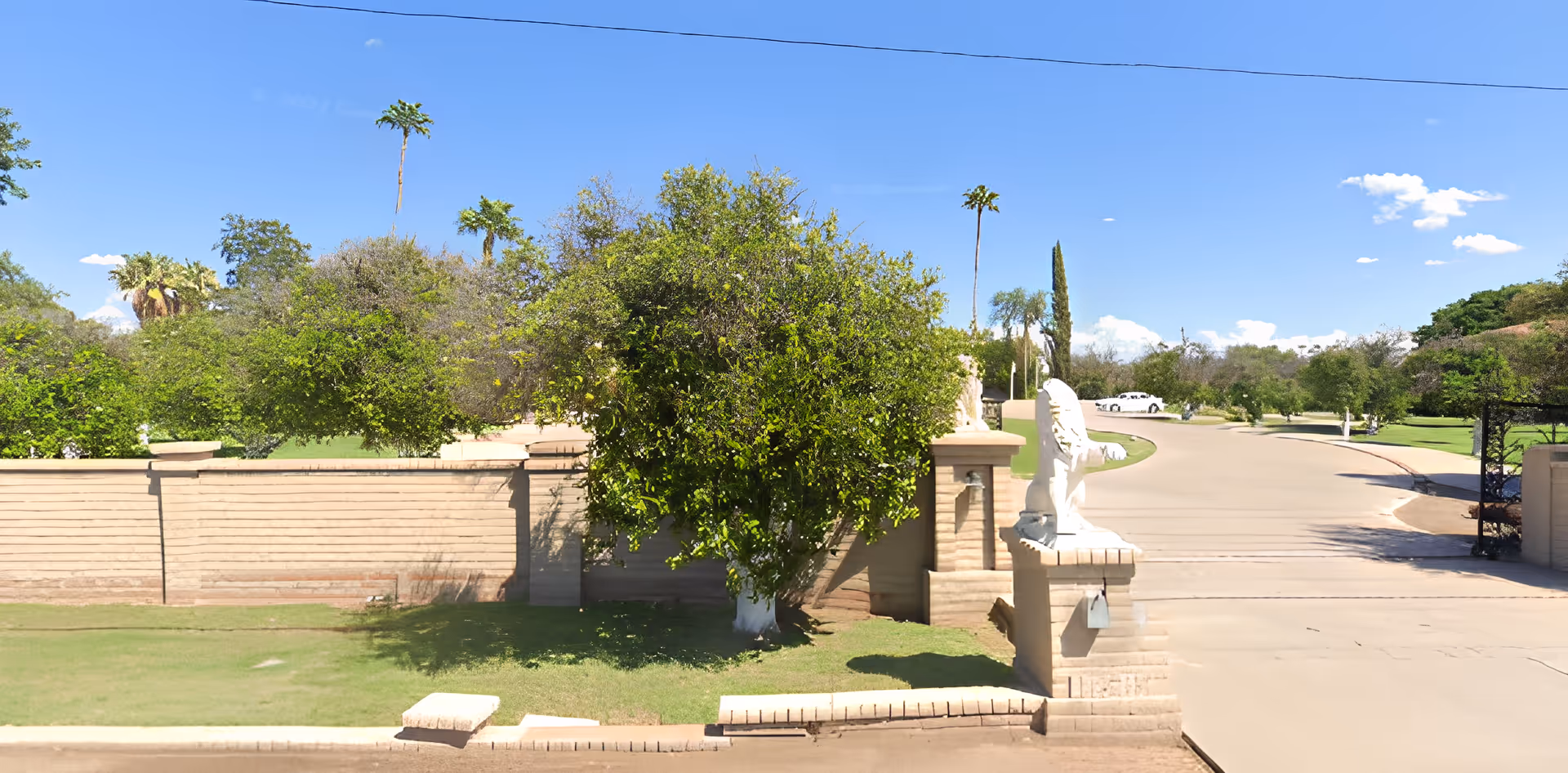 Entrance to a facility with a brick wall and two white lion statues on pillars flanking a driveway. There are green trees and a clear blue sky in the background.