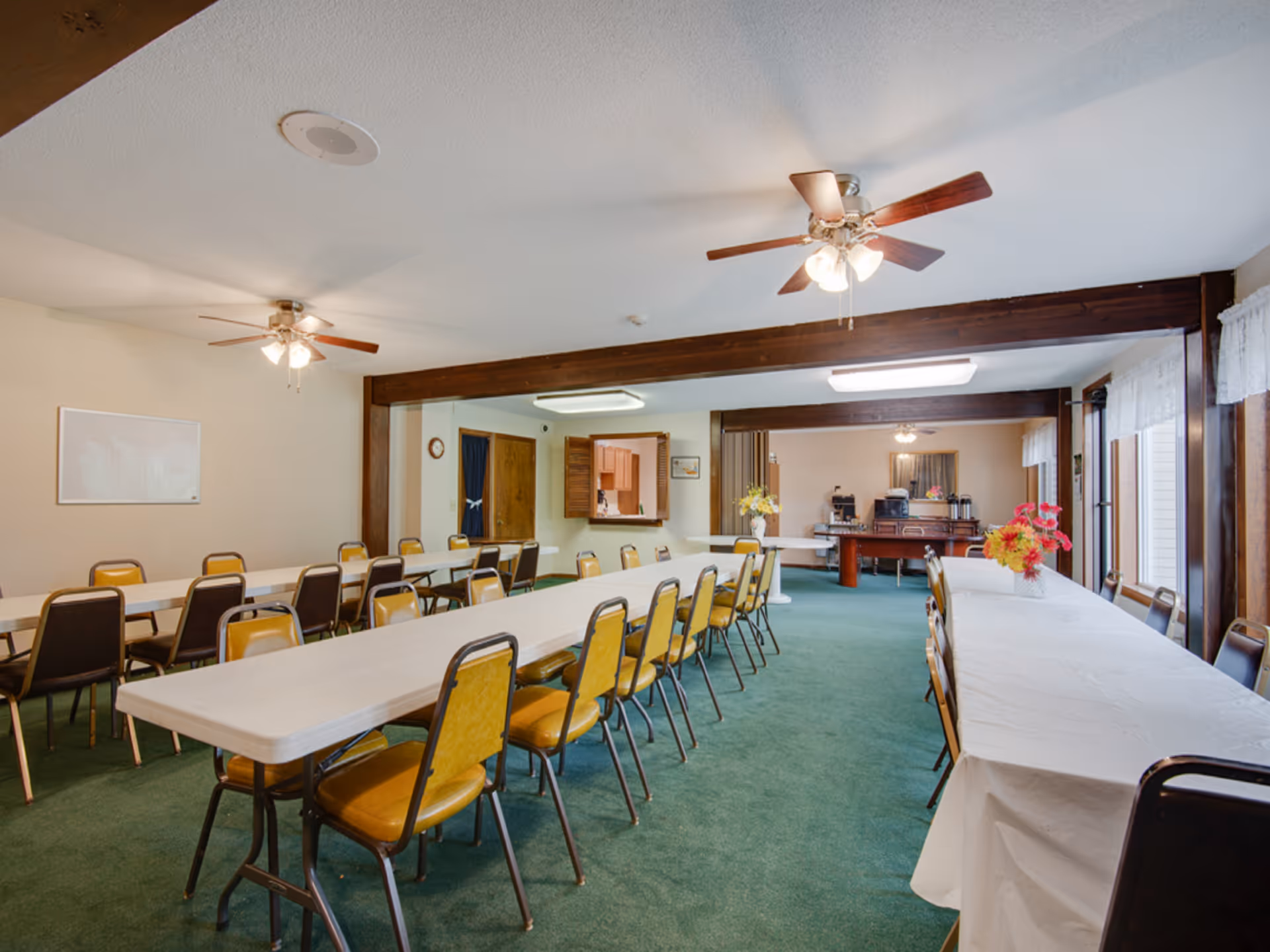 A large dining room with long tables covered with white tablecloths and surrounded by yellow and brown chairs. The room has green carpet, ceiling fans with lights, and large windows with white curtains. There are flower arrangements on the tables and a sideboard with coffee machines and other items in the background.