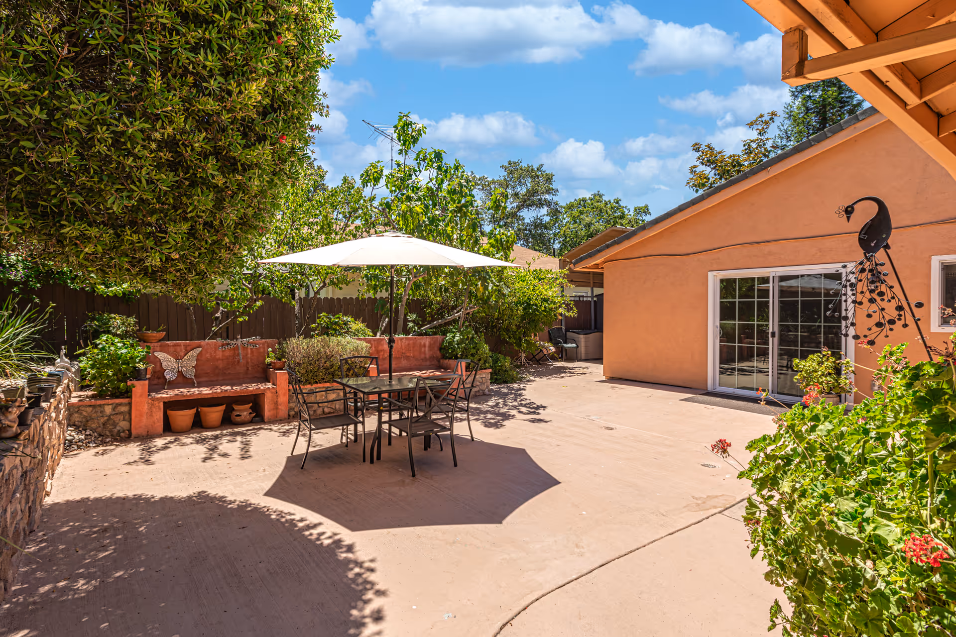 Outdoor patio area with a table and four chairs under a white umbrella, surrounded by greenery and plants. There is a peach-colored building with sliding glass doors and a decorative peacock sculpture on the wall. A stone bench with potted plants and a butterfly decoration is visible on the left side.