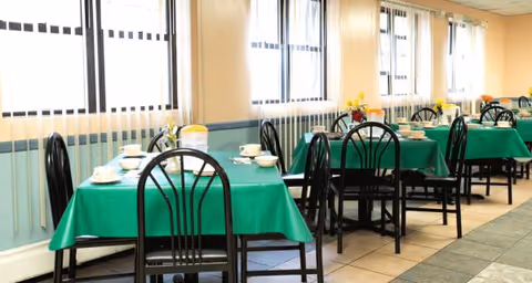 Bright dining room with green tablecloths, black chairs, and place settings along windows.