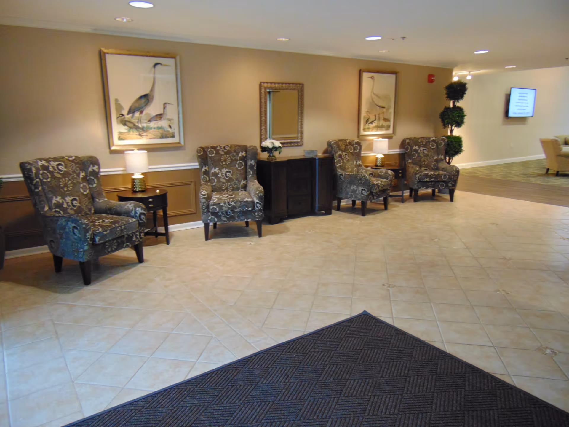 A waiting area with four patterned armchairs arranged along a beige wall. Between the chairs are small tables with lamps and a dark wooden cabinet with a mirror and a flower vase on top. Two framed bird illustrations hang on the wall above the chairs. The floor is tiled with a light beige pattern, and a dark entrance mat is visible in the foreground. In the background, there is a hallway with a TV mounted on the wall and a decorative topiary plant.