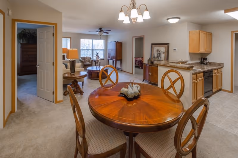 Interior view of a senior living facility showing a dining area with a round wooden table and four chairs, a kitchen with wooden cabinets and appliances, and a living room with sofas and a coffee table. There are doorways leading to other rooms, and the space is well-lit with ceiling lights and natural light from a window.
