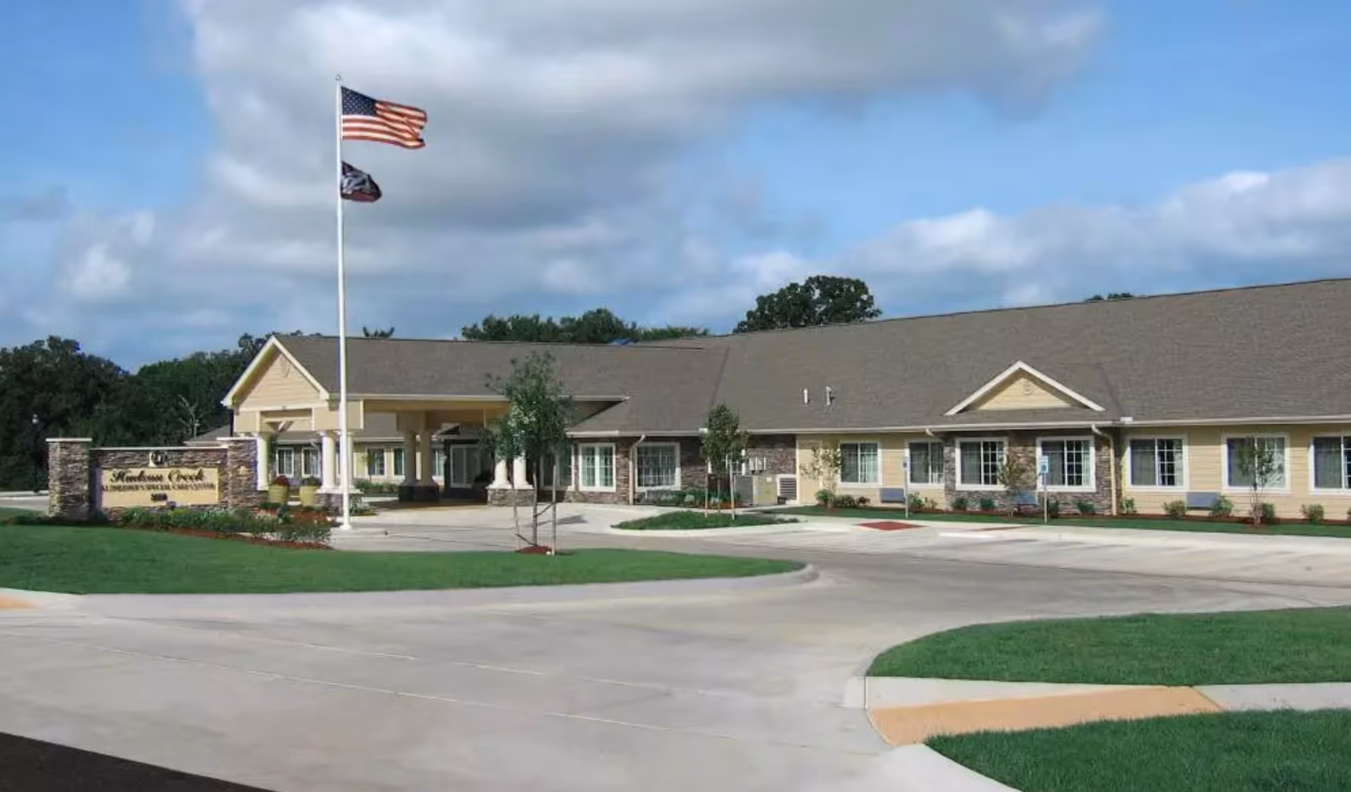 Front exterior of a single‑story beige senior living facility with an entrance canopy, American flags, landscaping, and a circular driveway.