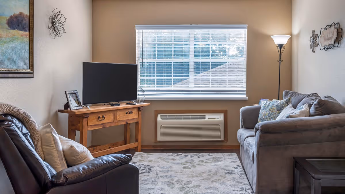 A cozy living room with a large window covered by white blinds. There is a wooden console table with a flat-screen TV and a framed photo on it. A gray couch with multiple pillows is on the right side, and a dark leather armchair with cushions is on the left. A floor lamp stands in the corner, and decorative wall art and a painting adorn the walls. A patterned area rug covers the wooden floor.