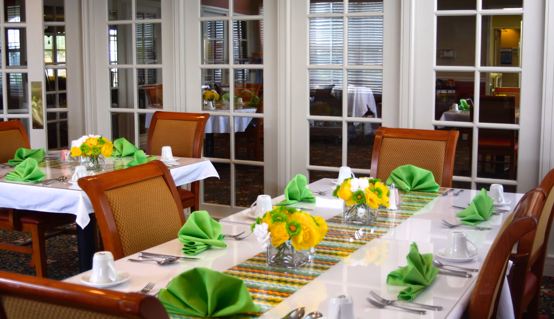 A dining room with tables set for a meal, featuring white tablecloths, green folded napkins, yellow and white flower centerpieces, and wooden chairs with cushioned seats. The background shows glass-paneled doors reflecting more tables and chairs.