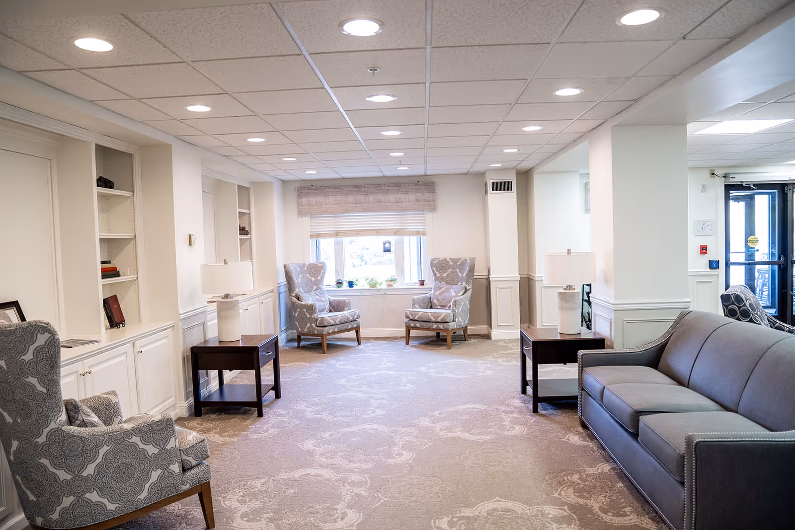 A well-lit common area with a gray sofa on the right, two patterned armchairs near a window at the back, and another patterned armchair on the left. There are two dark wooden side tables with white lamps on either side of the room. Built-in white shelves and cabinets line the left wall, and a glass door is visible on the right side near the entrance.