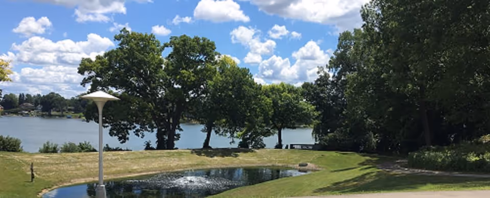 A scenic outdoor area at Lourdes Senior Community featuring a small pond with a fountain, green grass, several large trees, a walking path, and a lake in the background under a partly cloudy blue sky.