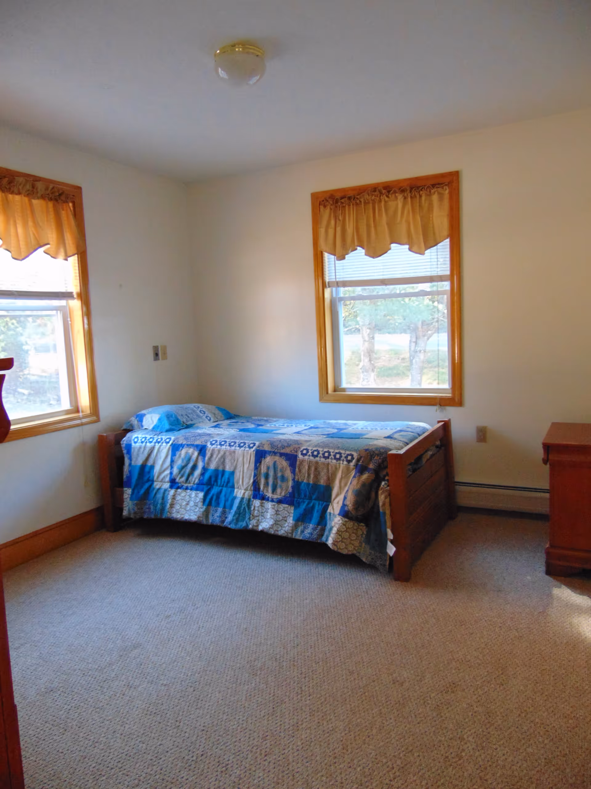 A simple bedroom with a single wooden bed covered with a blue and beige patchwork quilt. The room has two windows with beige curtains and wooden trim, beige carpet, and a wooden dresser on the right side.
