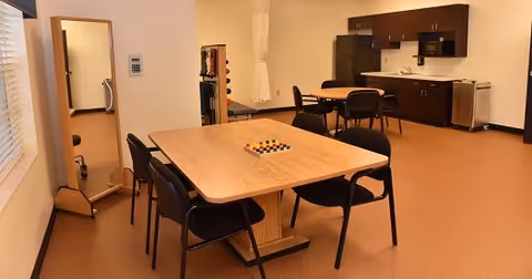 A room with a wooden table and four black chairs around it. On the table, there is a board game with colored pieces. In the background, there is a kitchenette with dark brown cabinets, a refrigerator, microwave, sink, and dishwasher. To the left, there is a full-length mirror and some exercise equipment partially visible. The room has beige walls and a brown floor.