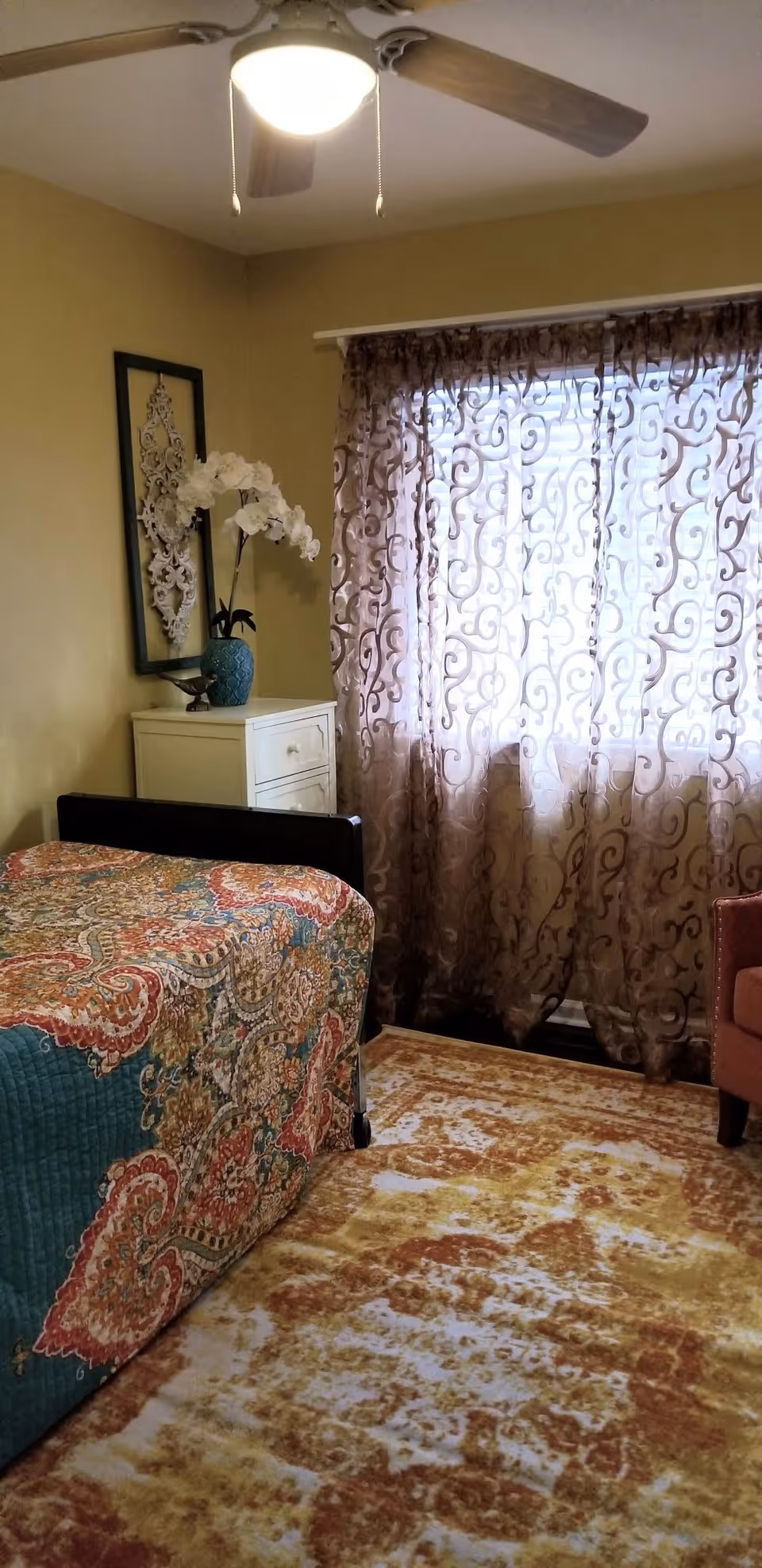 Sunlit bedroom with a patterned bedspread, ornate sheer curtains, a nightstand with a vase of flowers, a ceiling fan, and a patterned area rug.