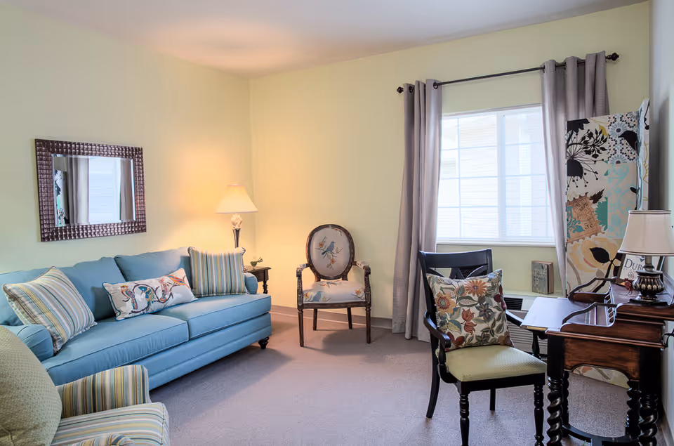 A cozy living room with a light blue sofa adorned with striped and patterned pillows, a floral upholstered armchair, and a wooden chair with a floral cushion. There is a wooden desk with a lamp and decorative items near a window with light gray curtains. A large mirror hangs on the pale yellow wall, and a floor lamp provides warm lighting.