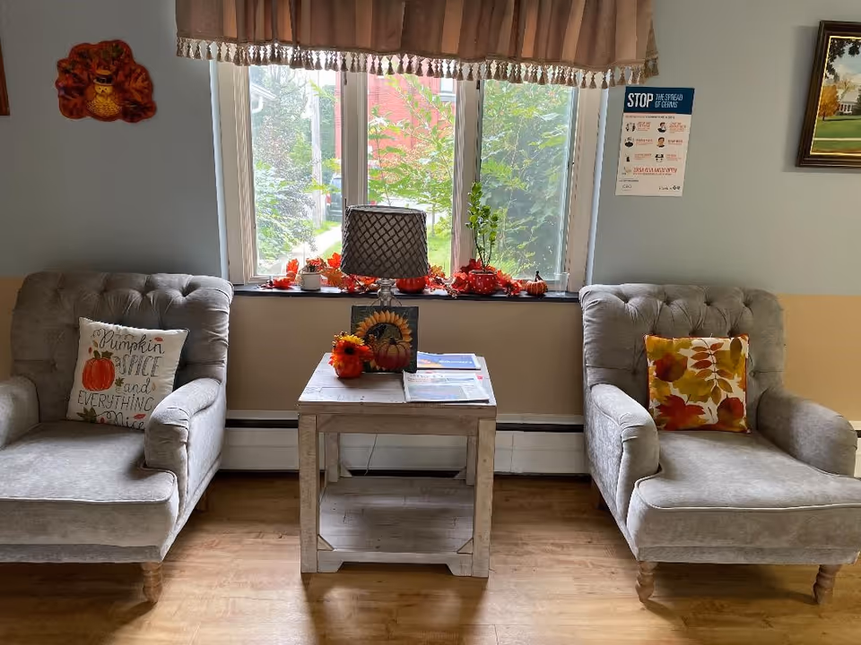 Two upholstered armchairs flank a small wooden table beneath a window decorated with fall accents.