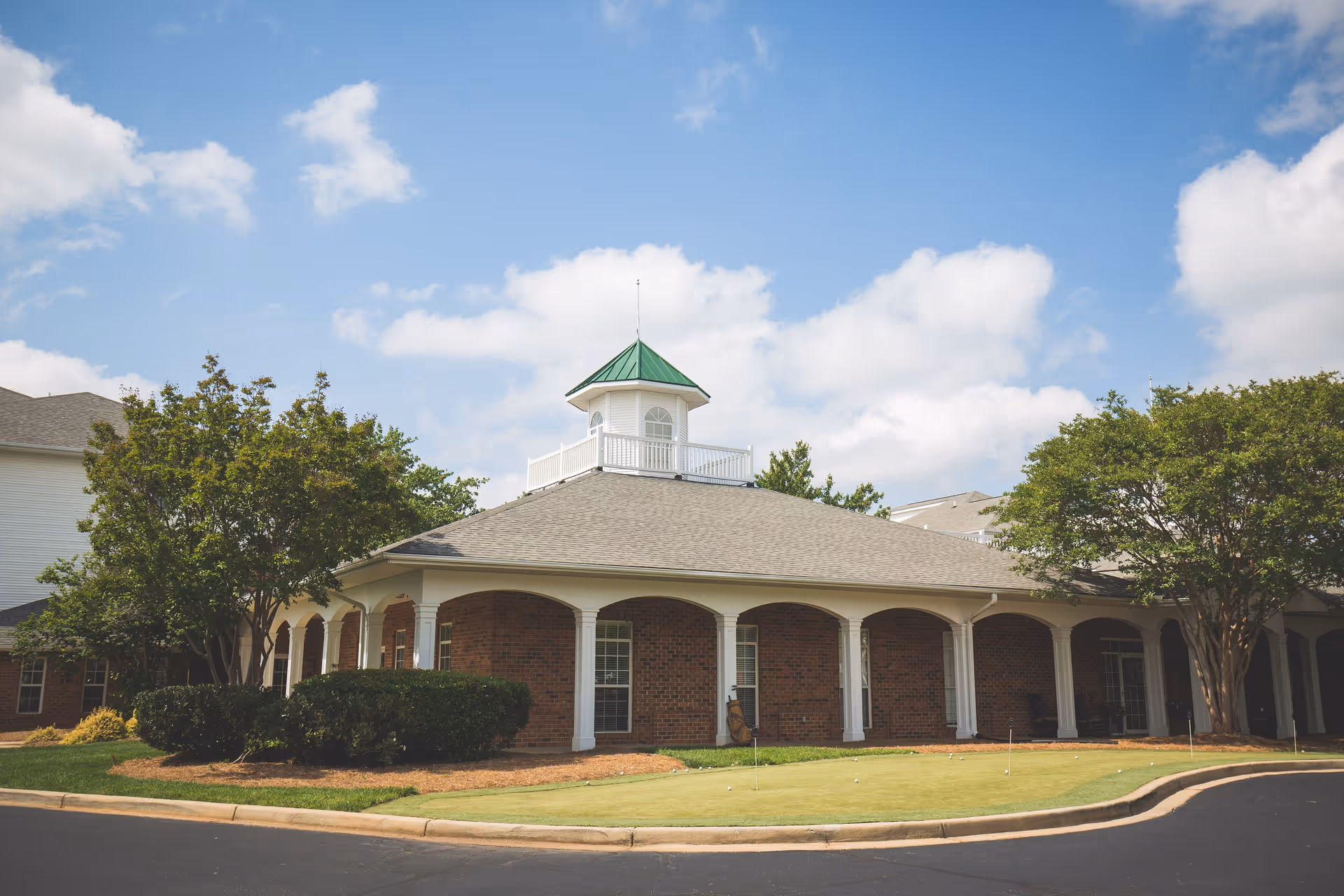 Exterior view of a brick building with white columns and a green-roofed cupola on top, surrounded by trees and bushes under a partly cloudy blue sky. There is a small putting green with golf balls and flags in front of the building.