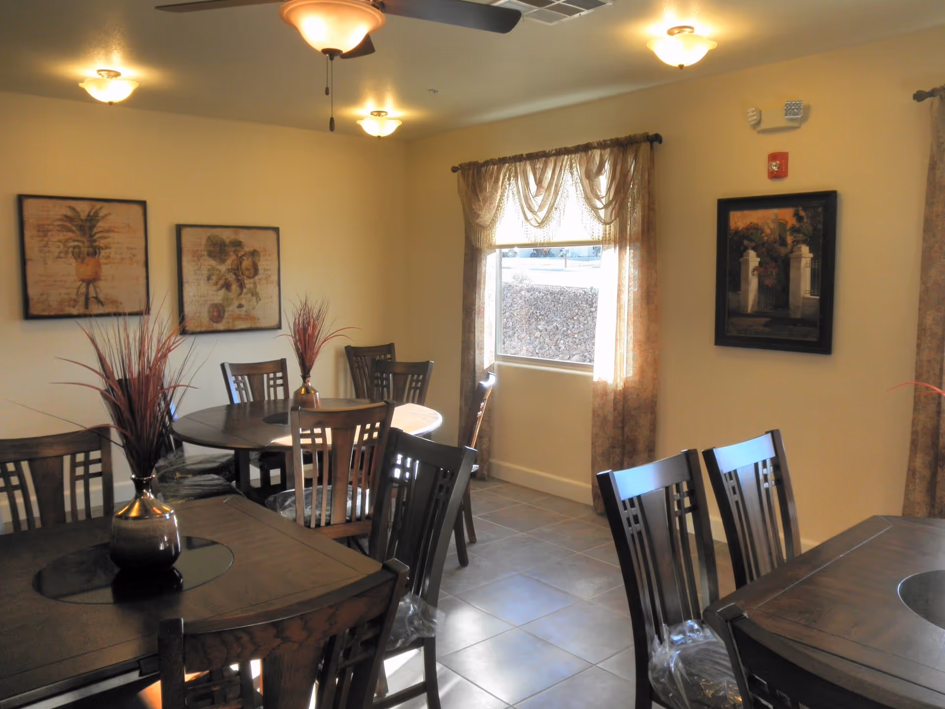 A dining room with multiple dark wooden tables and chairs. Each table has a decorative vase with red ornamental grass. The room has beige walls adorned with framed artwork and a window with sheer curtains letting in natural light. The floor is tiled, and ceiling lights provide additional illumination.
