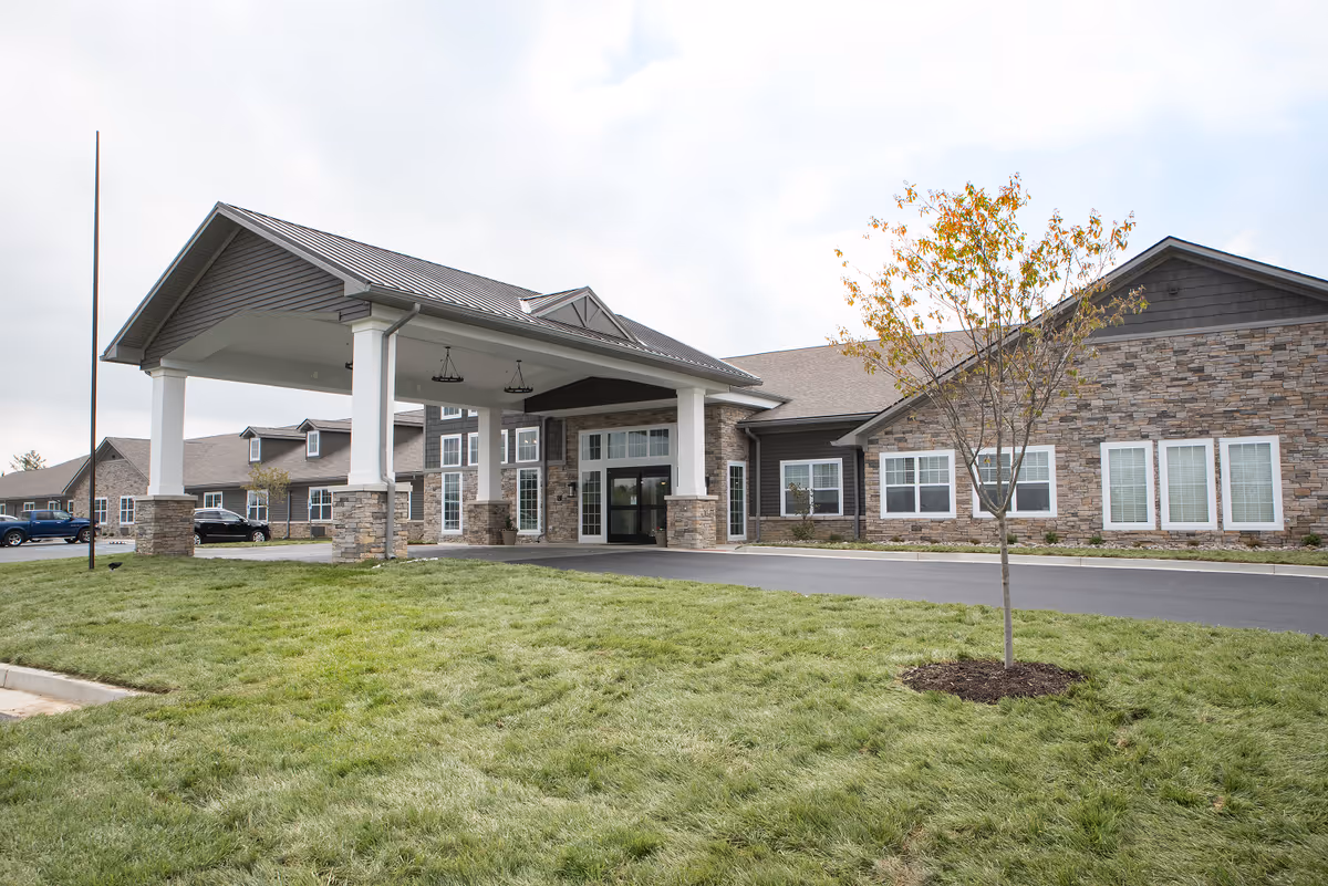 Exterior view of a senior living facility building with a covered entrance supported by white columns and stone bases. The building has a combination of stone and siding on the facade, multiple windows, and a small tree planted in the grassy area in front. Several cars are parked near the building.