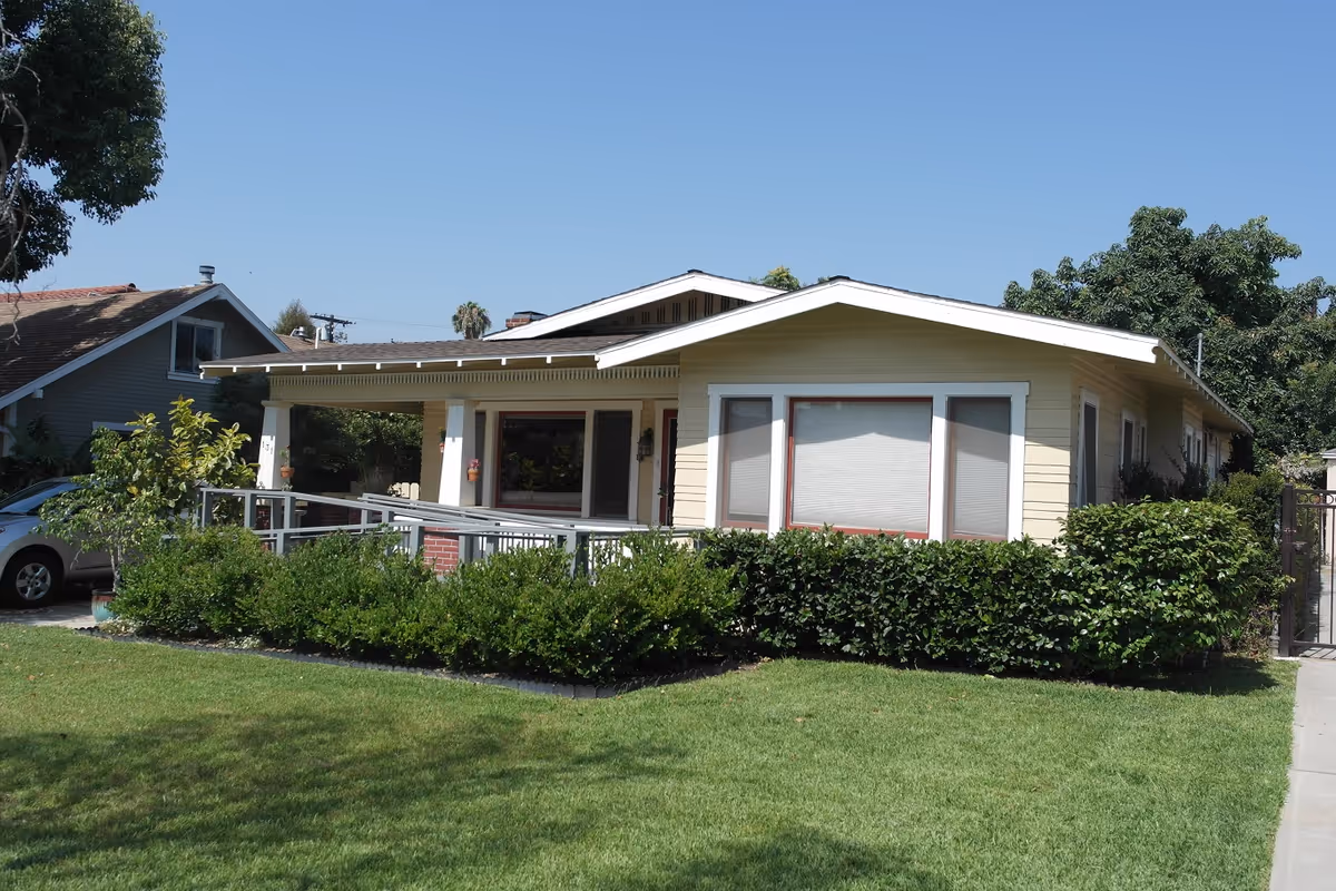 Single-story yellow house with white trim and a covered front porch with a wheelchair ramp, surrounded by green bushes and a well-maintained lawn under a clear blue sky.