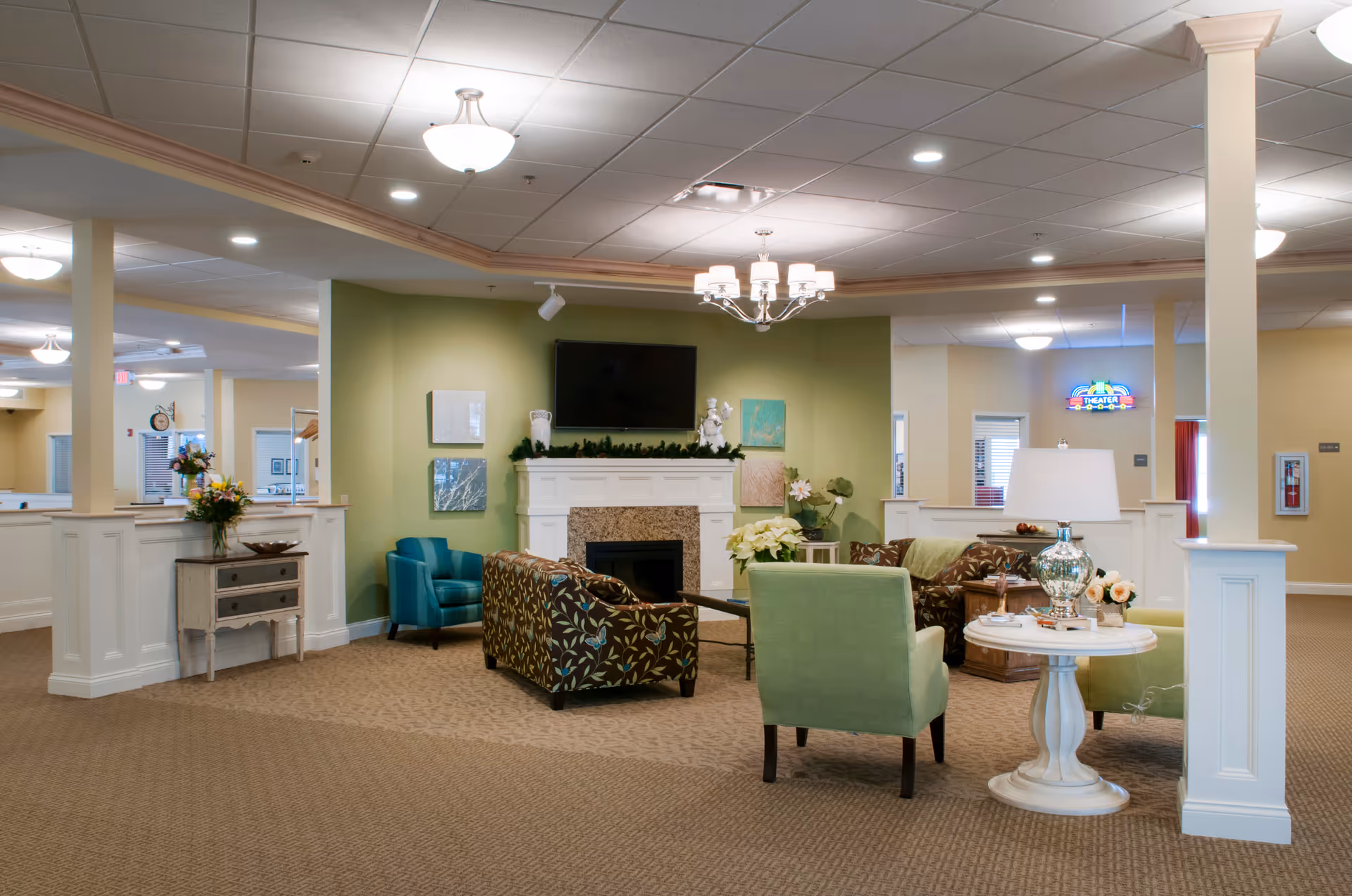 A cozy common area in a senior living facility featuring a fireplace with a TV mounted above it, surrounded by comfortable seating including patterned and solid-colored armchairs. The walls are painted in soft green and beige tones, with decorative artwork and plants. The space is well-lit with ceiling lights and a chandelier, and there is a small round table with a lamp and flowers in the foreground.