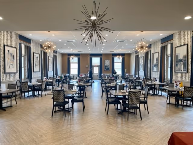 Spacious dining room with multiple tables and chairs, decorative starburst chandeliers, and framed artwork along the walls.
