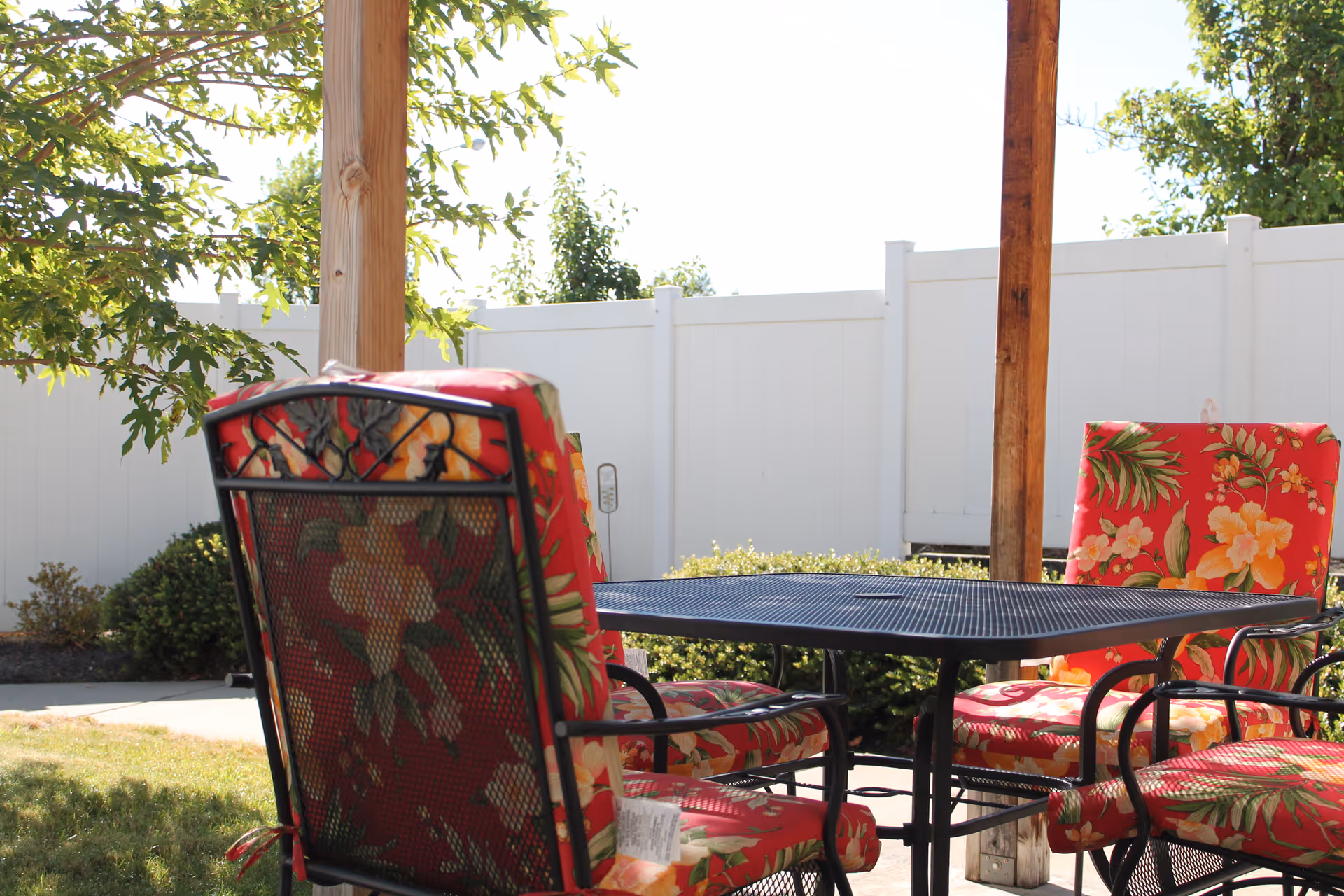Outdoor patio area with a black metal table and four chairs featuring red floral cushions, surrounded by greenery and a white fence in the background.