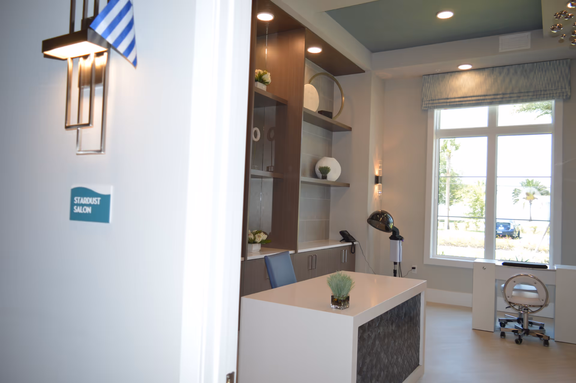 Interior view of a salon room named Stardust Salon with a white desk, a blue chair, decorative shelves with plants and ornaments, a hair dryer hood, and a large window with a view of trees and a parked car outside.