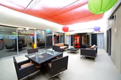 A modern indoor common area with a high ceiling featuring a red fabric canopy and colorful hanging lanterns. The space includes several dark wicker seating arrangements with cushions and a glass-top table. Large windows line one side of the room, and a vending machine is visible in the background.