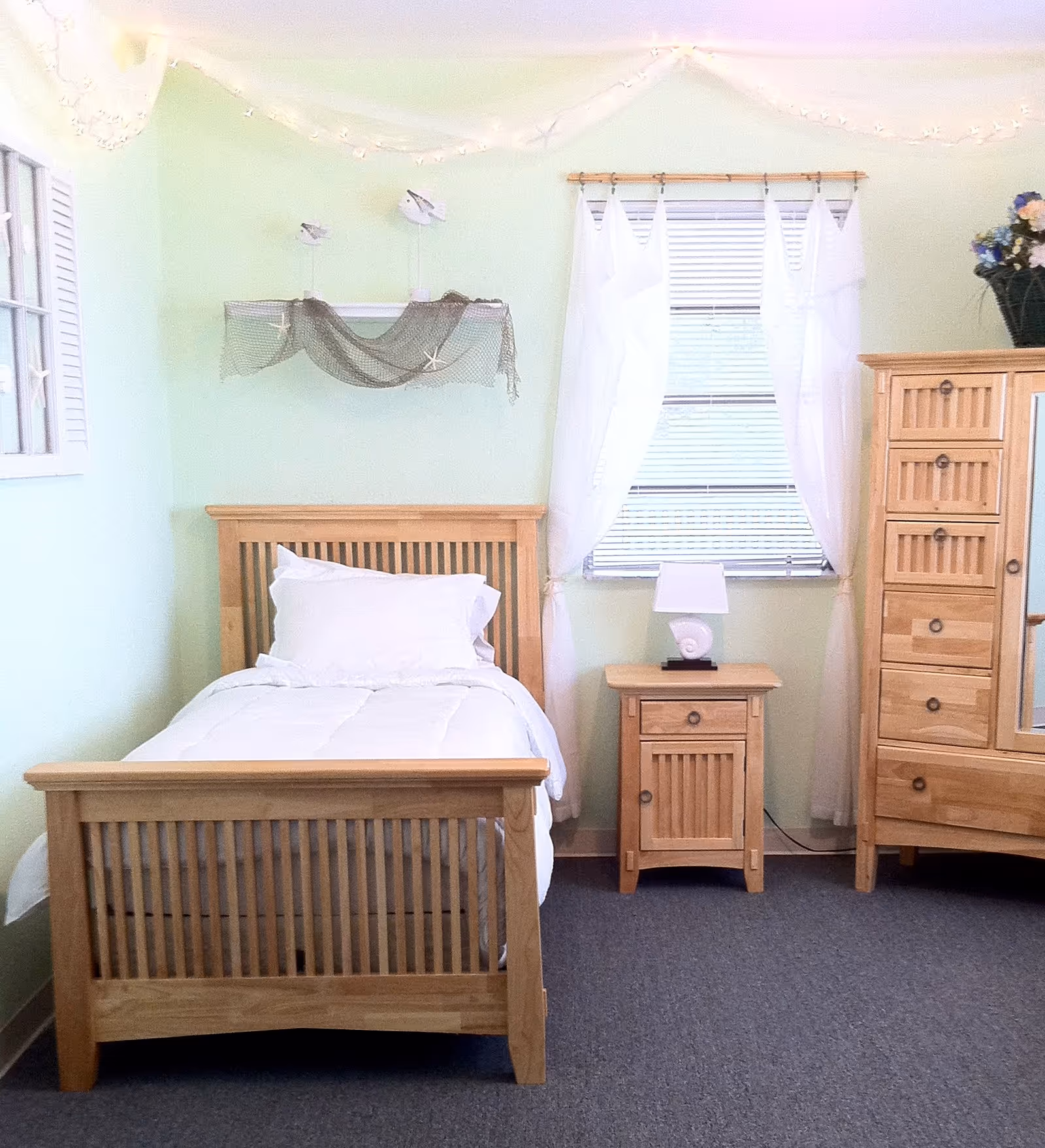 A neatly made single bed with white bedding and pillow in a room with light green walls. Next to the bed is a wooden nightstand with a white lamp on it. There is a window with white curtains behind the nightstand. To the right of the nightstand is a wooden dresser with multiple drawers and a mirror. The room has a carpeted floor and decorative string lights hanging near the ceiling.