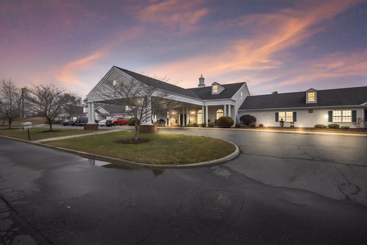 Exterior view of a senior living facility building at dusk with a covered entrance, lit windows, parked cars, and a partly cloudy sky with pink and purple hues.